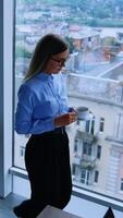 Lunch break in the modern office with beautiful view from window. Blonde female employee walking up to window with a cup of coffee. High angle view. Vertical video