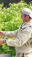 Caucasian male apiarist watching intently at the honey frame. Man turning in hands a frame coated heavily with bees. Green trees at backdrop. Vertical video