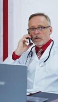 Senior physician consultation using mobile phone. Professional doctor in medical coat and stethoscope sitting in his cabinet and giving a conversation through the phone with a patient. Vertical video