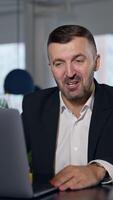 Business meeting via laptop. Businessman talking and smiling while talking before the laptop. Spacious office in blur at the background. Vertical video