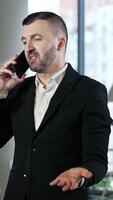 Businessman having a pleasant conversation on the mobile phone. Confident man has business talk on the phone. Big windows at the backdrop. Vertical video