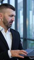 Caucasian man wearing black suit stands in the office and holds the laptop. Man with a personal computer in front of the panoramic window at work. Vertical video