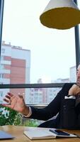 Business conversation on the laptop. Middle-aged entrepreneur sits at the table in front of the big panoramic windows. Cityscape at the backdrop. Vertical video
