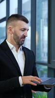 Male entrepreneur standing at the window and holding laptop in front of him. Business talk on the computer. Man talking and gesturing in front of notebook. Vertical video
