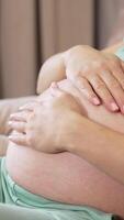 Caring future mother stroking her big tummy while resting on the sofa. Woman caressing her child in the big belly. Two little baby socks are on the armrest at he backdrop. Vertical video
