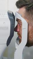 Patient man looks at device in ophthalmology clinic. Profile of patient's face looking into apparatus for eyes diagnostic in clinic. Close-up. Vertical video