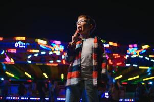 A cute caucasian boy eating a colorful lollipop standing against the background of a carousel in the evening at an amusement park or circus photo