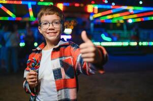 A cute caucasian boy eating a colorful lollipop standing against the background of a carousel in the evening at an amusement park or circus photo