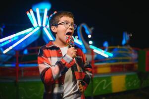 A cute caucasian boy eating a colorful lollipop standing against the background of a carousel in the evening at an amusement park or circus photo
