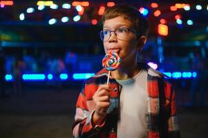 A cute caucasian boy eating a colorful lollipop standing against the background of a carousel in the evening at an amusement park or circus photo
