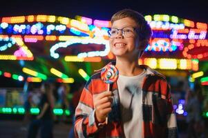 A cute caucasian boy eating a colorful lollipop standing against the background of a carousel in the evening at an amusement park or circus photo
