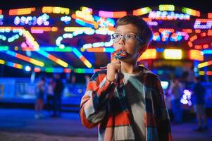 A cute caucasian boy eating a colorful lollipop standing against the background of a carousel in the evening at an amusement park or circus photo