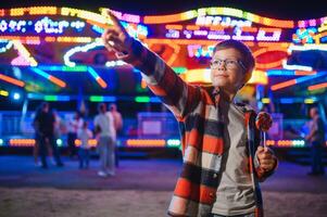 A cute caucasian boy eating a colorful lollipop standing against the background of a carousel in the evening at an amusement park or circus photo