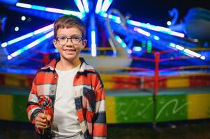 A cute caucasian boy eating a colorful lollipop standing against the background of a carousel in the evening at an amusement park or circus photo