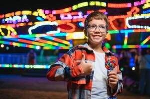 A cute caucasian boy eating a colorful lollipop standing against the background of a carousel in the evening at an amusement park or circus photo