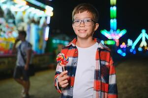 A cute caucasian boy eating a colorful lollipop standing against the background of a carousel in the evening at an amusement park or circus photo