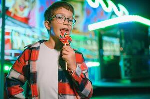 A cute caucasian boy eating a colorful lollipop standing against the background of a carousel in the evening at an amusement park or circus photo