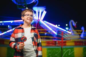 A cute caucasian boy eating a colorful lollipop standing against the background of a carousel in the evening at an amusement park or circus photo