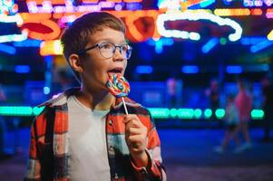 A cute caucasian boy eating a colorful lollipop standing against the background of a carousel in the evening at an amusement park or circus photo