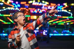 A cute caucasian boy eating a colorful lollipop standing against the background of a carousel in the evening at an amusement park or circus photo