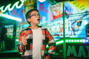 A cute caucasian boy eating a colorful lollipop standing against the background of a carousel in the evening at an amusement park or circus photo