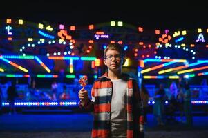 A cute caucasian boy eating a colorful lollipop standing against the background of a carousel in the evening at an amusement park or circus photo
