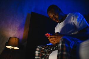 Low angle portrait of adult African-American man using smartphone while sitting on bed at night. Problems with insomnia. photo