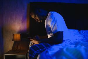 Low angle portrait of adult African-American man using smartphone while sitting on bed at night. Problems with insomnia. photo