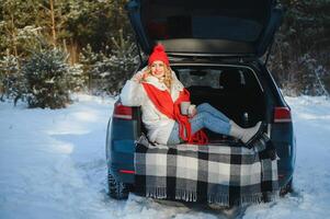 young woman sitting in car trunk drinking warm tea and takes a selfie at winter snowed day. photo