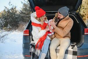 Smiling couple with dog sitting in open SUV car trunk in snowy forest. Enjoying each other in active winter holidays. photo