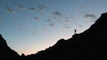 Silhouette Amazing view of dark mountain range with orange blue skyline, happy male hiker man on a stone on a summit raising hands up with delight, overwhelmed with emotions. High quality 4k footage video