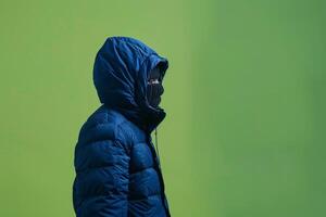 A lone figure wrapped in a dark coat walks against a bright green backdrop on a chilly day photo