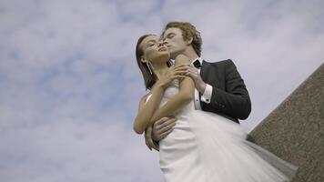 Bottom view of stylish caring bride and groom on blue sky background with clouds. Action. Man in black and white suit kissing woman cheek in white dress. photo