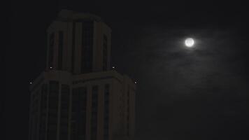 Night cityscape with a high building on dark sky background. Stock footage. Full moon on dark cloudy sky. photo