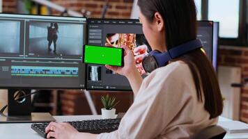 Asian female editor working from home for freelance work with green screen, using smartphone and dual monitors for mixing software. Creative workspace equipped with tech tools. Camera B. video