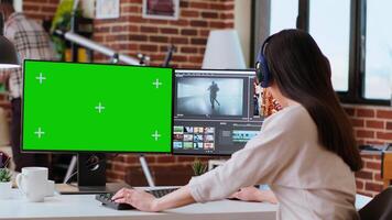 Professional asian woman working on a media project next to green screen in a modern space, using dual monitors and advanced editing software. Multitasking in post production. Camera A. video