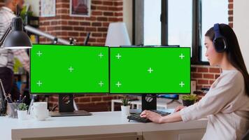 Asian freelancer working on her tasks with green screen on pc display, listening to music on headset while she does research at home. Remote working on freelance job, multitasking. Camera B. video