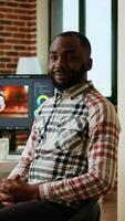 Vertical African american male filmmaker works with creative editing software, mixing and color grading clips. Living room workspace features post production tools and dual monitors. Camera A. video