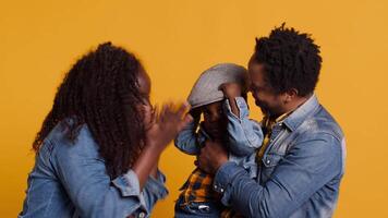 African american joyful family posing together against yellow background, happy couple hugging their little toddler. Smiling young mother and father showing affection to their kid. Camera A. video