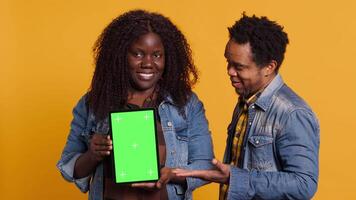 African american couple pointing at a tablet with isolated green screen display, smiling at each other against yellow background. Cheerful affectionate husband and wife posing. Camera A. video