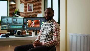 African american male filmmaker works with creative editing software, mixing and color grading clips. Living room workspace features post production tools and dual monitors. Camera A. video