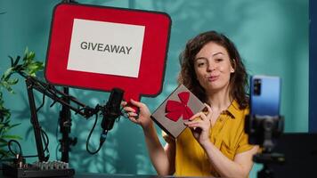 Smiling woman holding empty copy space sign, excited to do giveaway for subscribers. Media star happy to offer packed gift with ribbon to viewers, holding cardboard bubble speech cutout, camera A video