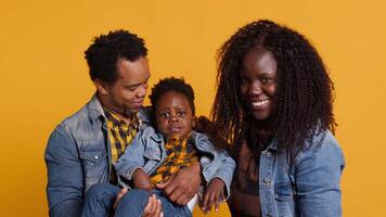 African american family smiling against yellow background in studio, posing with love and sincere gratitude. Positive optimistic mom and dad holding their baby boy, enjoying time together. Camera A. video