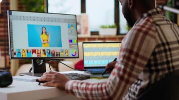 African american male designer edits images on dual monitors, computer setup in a creative office. Ideal workspace for digital media professional focused on photo retouching. Camera B. video
