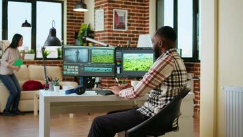 Male editor works from modern home office in the living room, multitasking with a dual monitor setup and editing software. Finishing post production tasks for media production. Camera A. video