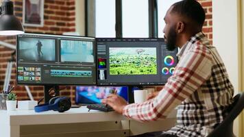 African american man editor working in production industry, does color grading and editing a film project on dual monitors. Showing the creative process with tech tools. Camera B. video