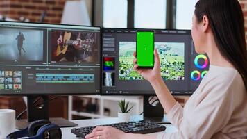 Professional asian woman working on a media project next to green screen in a modern space, using dual monitors and advanced editing software. Multitasking in post production. Camera A. video