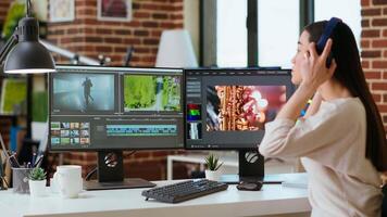 Asian filmmaker woman putting headphones on and working on production at a home office, mixing clips. Office setup with a software perfect for color grading in post production. Camera B. video