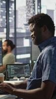 Vertical Employee with grimace on face stretches to relieve muscle strain while conducting code review to fix bugs. Fatigued staff member in office suffers back pain from bad posture, optimizing code, camera B video