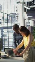 Vertical African american supervisor looking at laptop screen with employee, checking on finished project task. Company management executive and woman analyzing data on notebook, camera B video